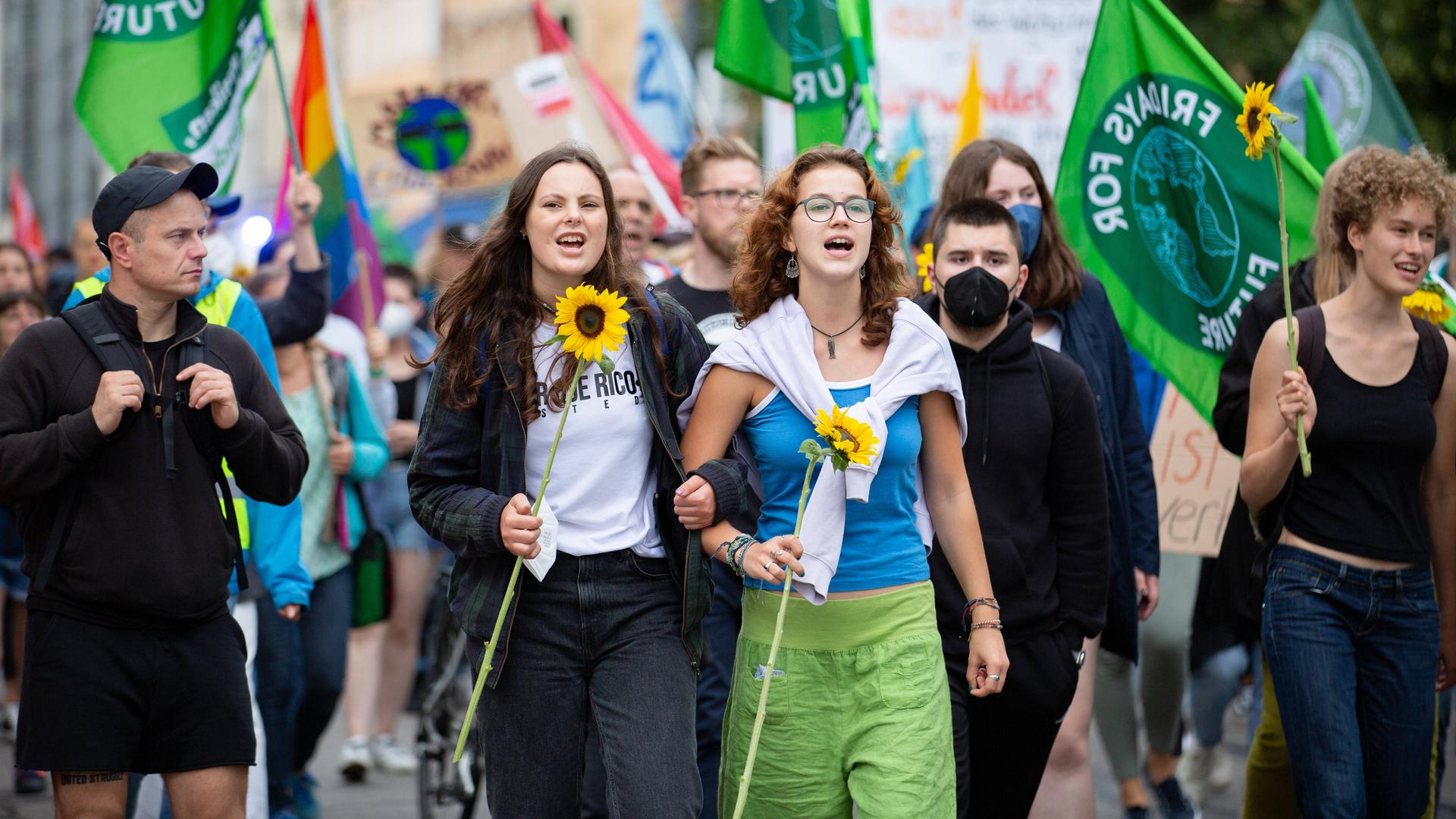 Zwei junge Frauen mit Sonnenblumen auf einer Demo. Im Hintergrund viele Fridays for Future-Fahnen.