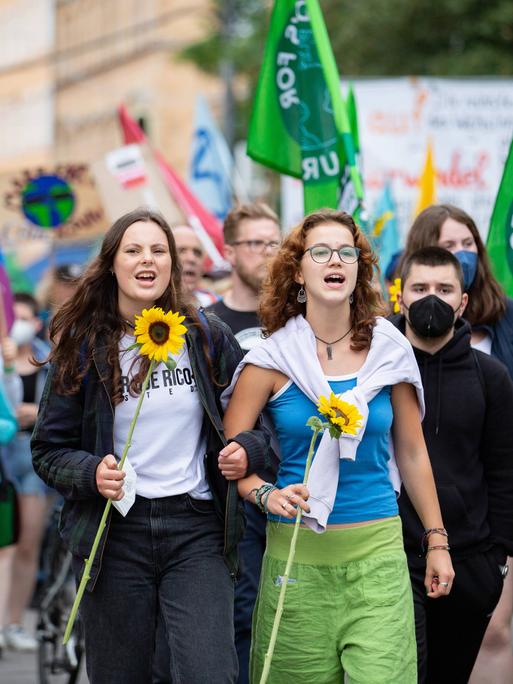 Zwei junge Frauen mit Sonnenblumen auf einer Demo. Im Hintergrund viele Fridays for Future-Fahnen.