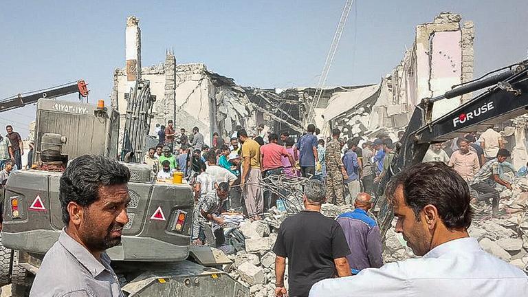 In der Ruine der Schule auf den Trümmern stehen viele Menschen. Im Vordergrund zwei weitere Personen und ein Bagger.