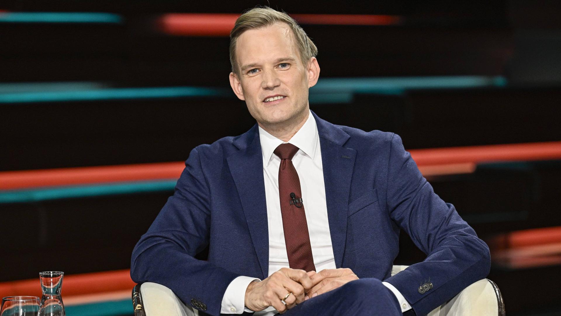 Hendrik Streeck sitzt in einem TV-Studio mit einem blauen Anzug in einem Sessel und schaut geradeaus in die Kamera. Hendrik Streeck sitzt in einem TV-Studio mit einem blauen Anzug in einem Sessel und schaut geradeaus in die Kamera.
