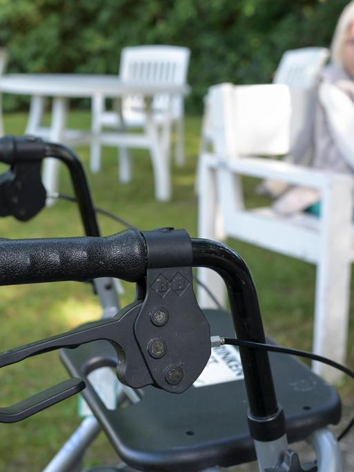 Ein Rollator im Vordergrund des Bildes, im Hintergrund, unscharf eine ältere Dame, vermutlich die Person, die auf diese Gehilfe angewiesen ist Ein Rollator im Vordergrund des Bildes, im Hintergrund, unscharf eine ältere Dame, vermutlich die Person, die auf diese Gehilfe angewiesen ist
