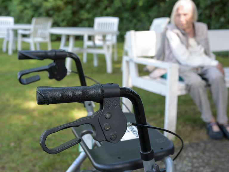 Ein Rollator im Vordergrund des Bildes, im Hintergrund, unscharf eine ältere Dame, vermutlich die Person, die auf diese Gehilfe angewiesen ist Ein Rollator im Vordergrund des Bildes, im Hintergrund, unscharf eine ältere Dame, vermutlich die Person, die auf diese Gehilfe angewiesen ist