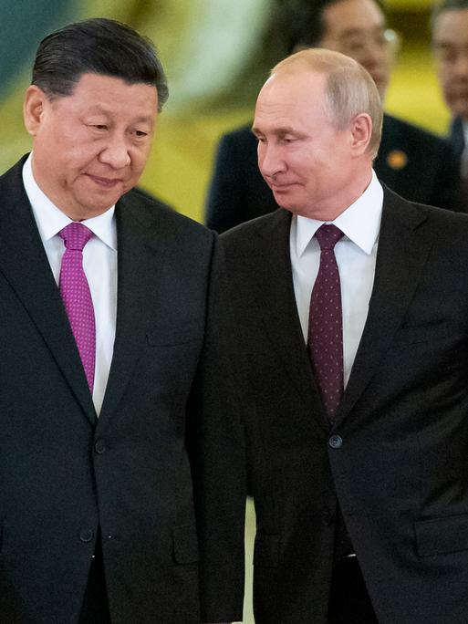 Chinas Präsident Xi Jinping und Russlands Staatschef Wladimir Putin gehen in schwarzen Anzügen gekleidet nebeneinander her, hinter ihnen weitere Männer in schwarzen Anzügen.