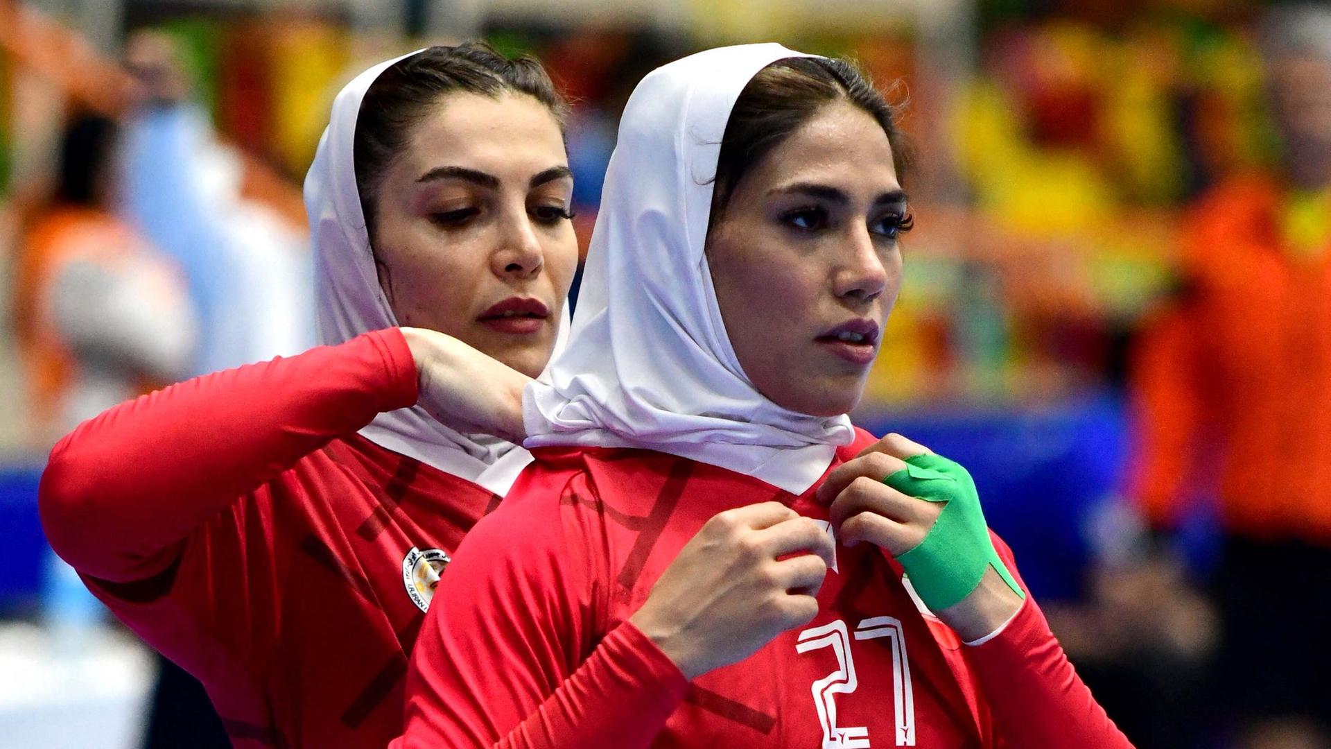 Die zwei Spielerinnen tragen rote Trikots und weiße Kopftücher: Eine bindet der anderen das Tuch hinten fest. 