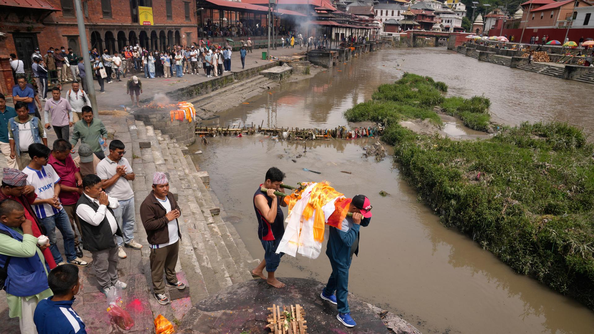 Angehörige tragen symbolisch einen Sarg einer vermissten Person am Fluss Bagmati entlang. Entlang des Flusses stehen noch Hunderte Menschen.