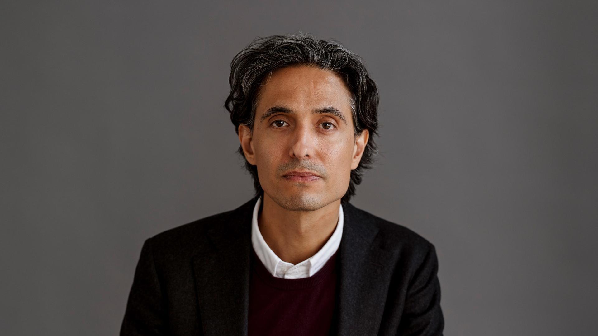 Der schwedische Schriftsteller Jonas Hassen Khemiri trägt einen schwarzen Anzug und sitzt in einem Fotostudio mit grauem Hintergrund