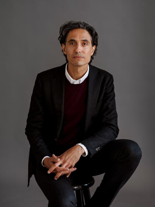 Der schwedische Schriftsteller Jonas Hassen Khemiri trägt einen schwarzen Anzug und sitzt in einem Fotostudio mit grauem Hintergrund Der schwedische Schriftsteller Jonas Hassen Khemiri trägt einen schwarzen Anzug und sitzt in einem Fotostudio mit grauem Hintergrund