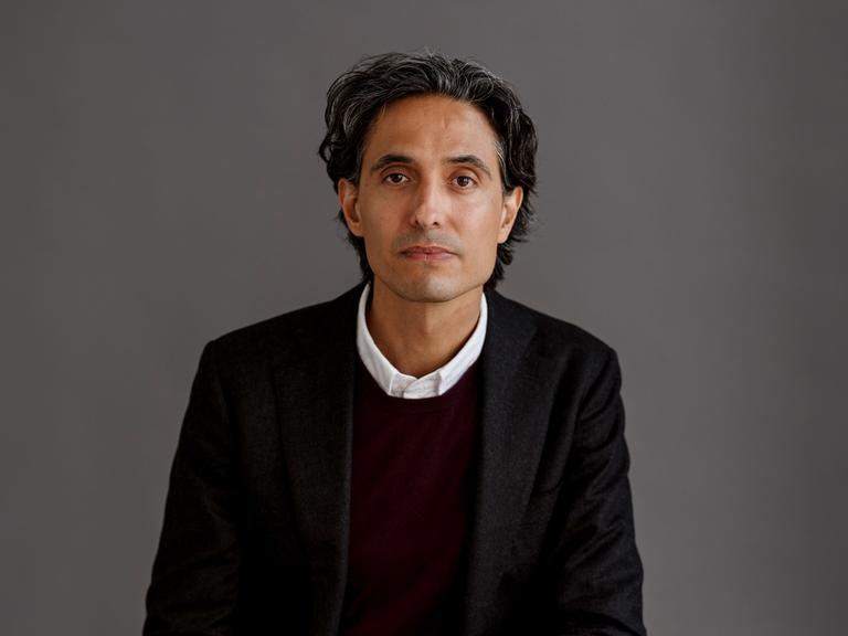 Der schwedische Schriftsteller Jonas Hassen Khemiri trägt einen schwarzen Anzug und sitzt in einem Fotostudio mit grauem Hintergrund Der schwedische Schriftsteller Jonas Hassen Khemiri trägt einen schwarzen Anzug und sitzt in einem Fotostudio mit grauem Hintergrund