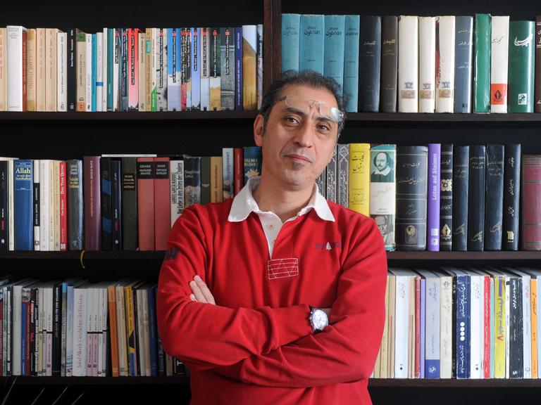 Madjid Mohit vom Sujet Verlag steht vor einem Regal mit einigen der von ihm verlegten Bücher.