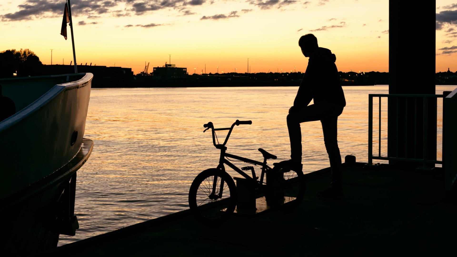 Die Silhouette eines jungen Mannes mit seinem Fahrrad auf einem Steg im warmen Abendlicht