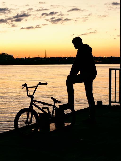 Die Silhouette eines jungen Mannes mit seinem Fahrrad auf einem Steg im warmen Abendlicht Die Silhouette eines jungen Mannes mit seinem Fahrrad auf einem Steg im warmen Abendlicht