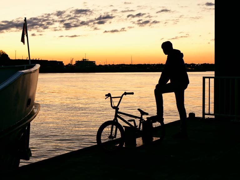 Die Silhouette eines jungen Mannes mit seinem Fahrrad auf einem Steg im warmen Abendlicht Die Silhouette eines jungen Mannes mit seinem Fahrrad auf einem Steg im warmen Abendlicht