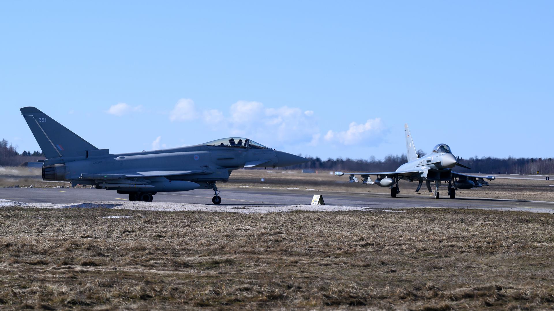 Zwei hellgrau lackierte Kampfjets stehen unter blauem Himmel auf einem Rollfeld.