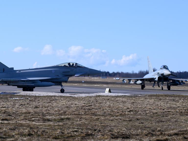 Zwei hellgrau lackierte Kampfjets stehen unter blauem Himmel auf einem Rollfeld.
