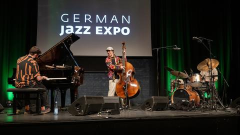 Der Bassist Nils Kugelmann steht im Zentrum einer Bühne auf der Messe jazzahead am Bass, neben ihm ein Flügel und ein Schlagzeug. Der Bassist Nils Kugelmann steht im Zentrum einer Bühne auf der Messe jazzahead am Bass, neben ihm ein Flügel und ein Schlagzeug.