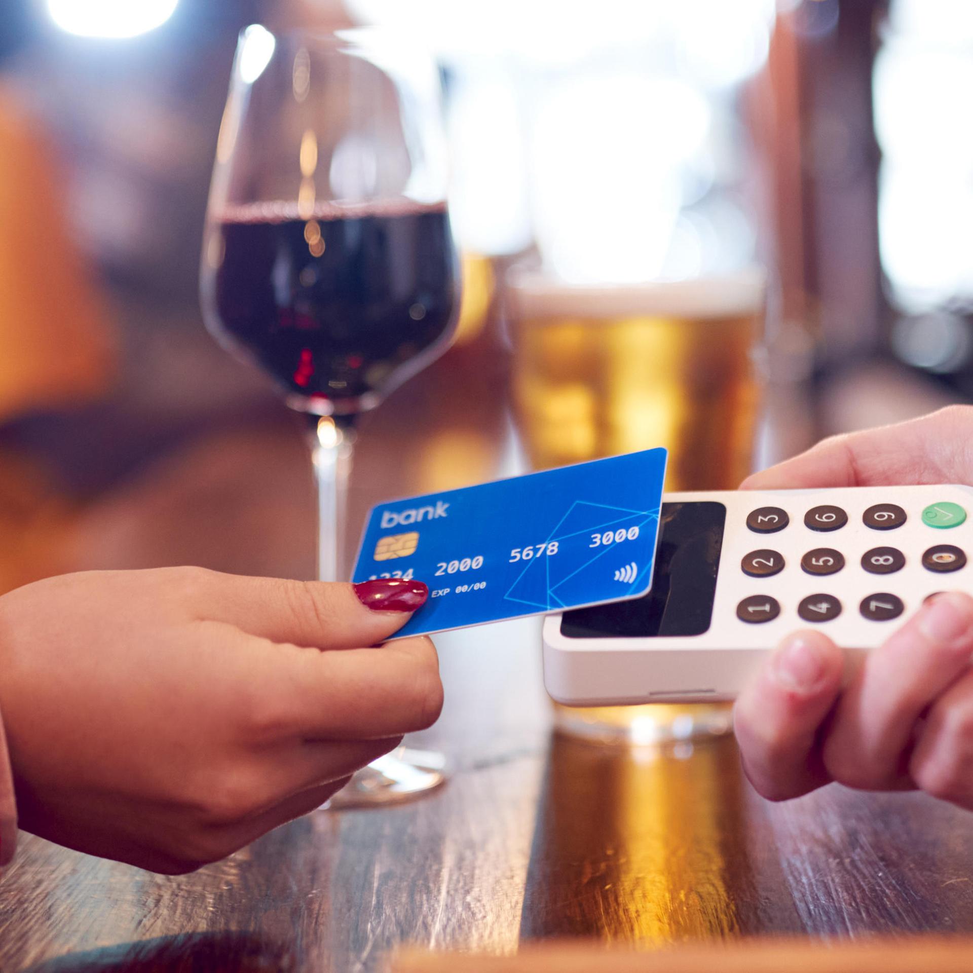 Eine Frauenhand mit dunkelrot lackierten Nägeln legt ihre Kreditkarte auf ein Kartenlesegerät. Im Hintergrund ist ein Glas mit Rotwein zu sehen.