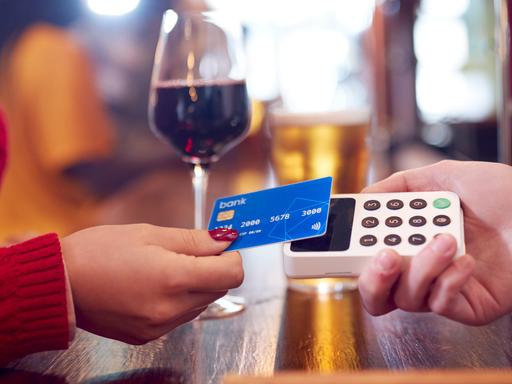 Eine Frauenhand mit dunkelrot lackierten Nägeln legt ihre Kreditkarte auf ein Kartenlesegerät. Im Hintergrund ist ein Glas mit Rotwein zu sehen.