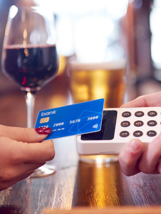 Eine Frauenhand mit dunkelrot lackierten Nägeln legt ihre Kreditkarte auf ein Kartenlesegerät. Im Hintergrund ist ein Glas mit Rotwein zu sehen.