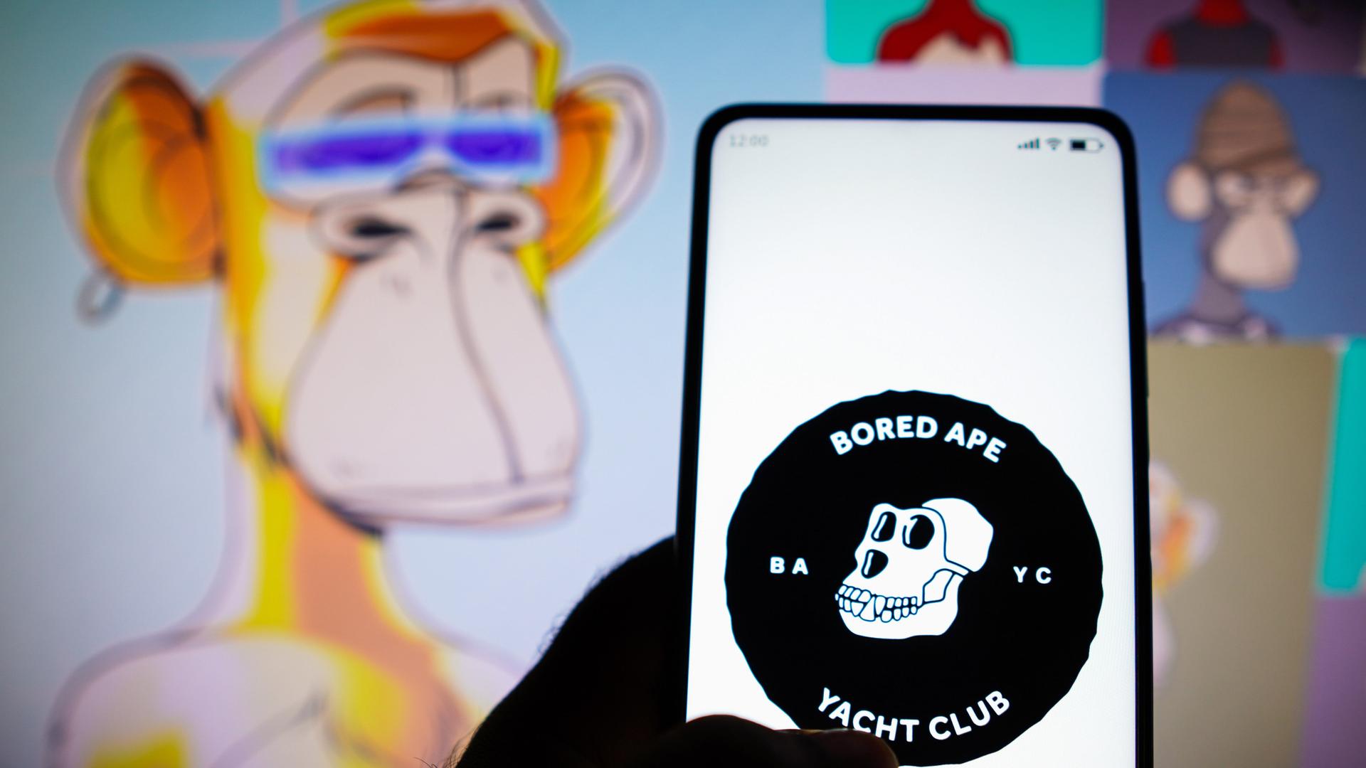 Vor einigen Grafiken mit den gelangweilten Affen hält eine Hand ein Smartphone auf dem das Logo des "Bored Ape Yacht Club" zu sehen ist: ein Affenschädel mit dem Namen und der Abkürzung BAYC.