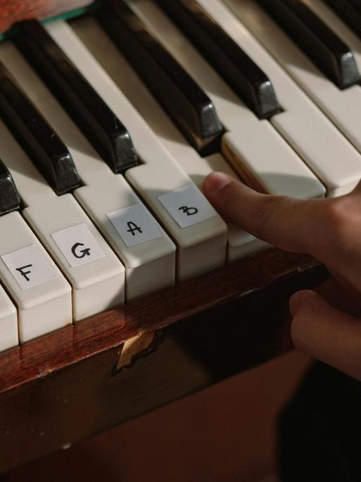 Eine Klaviatur mit aufgeklebten Notennamen auf den entsprechenden Tasten. Dazu eine spielende Hand.