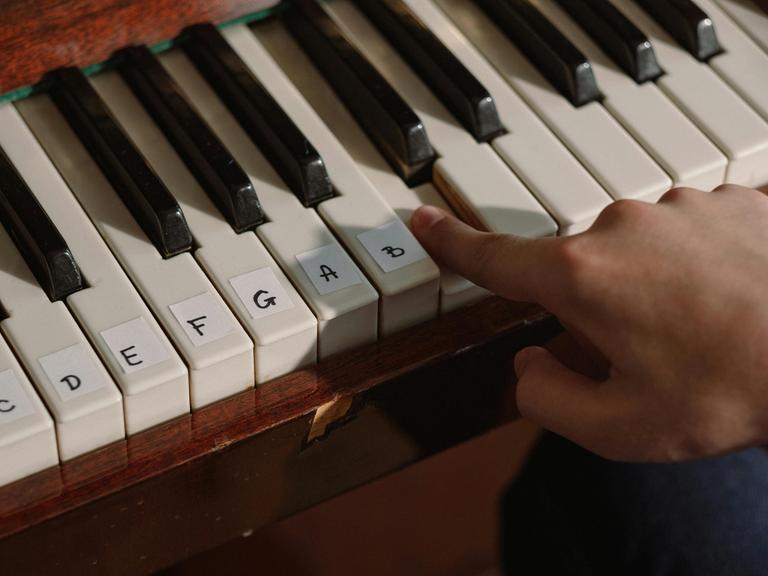 Eine Klaviatur mit aufgeklebten Notennamen auf den entsprechenden Tasten. Dazu eine spielende Hand.