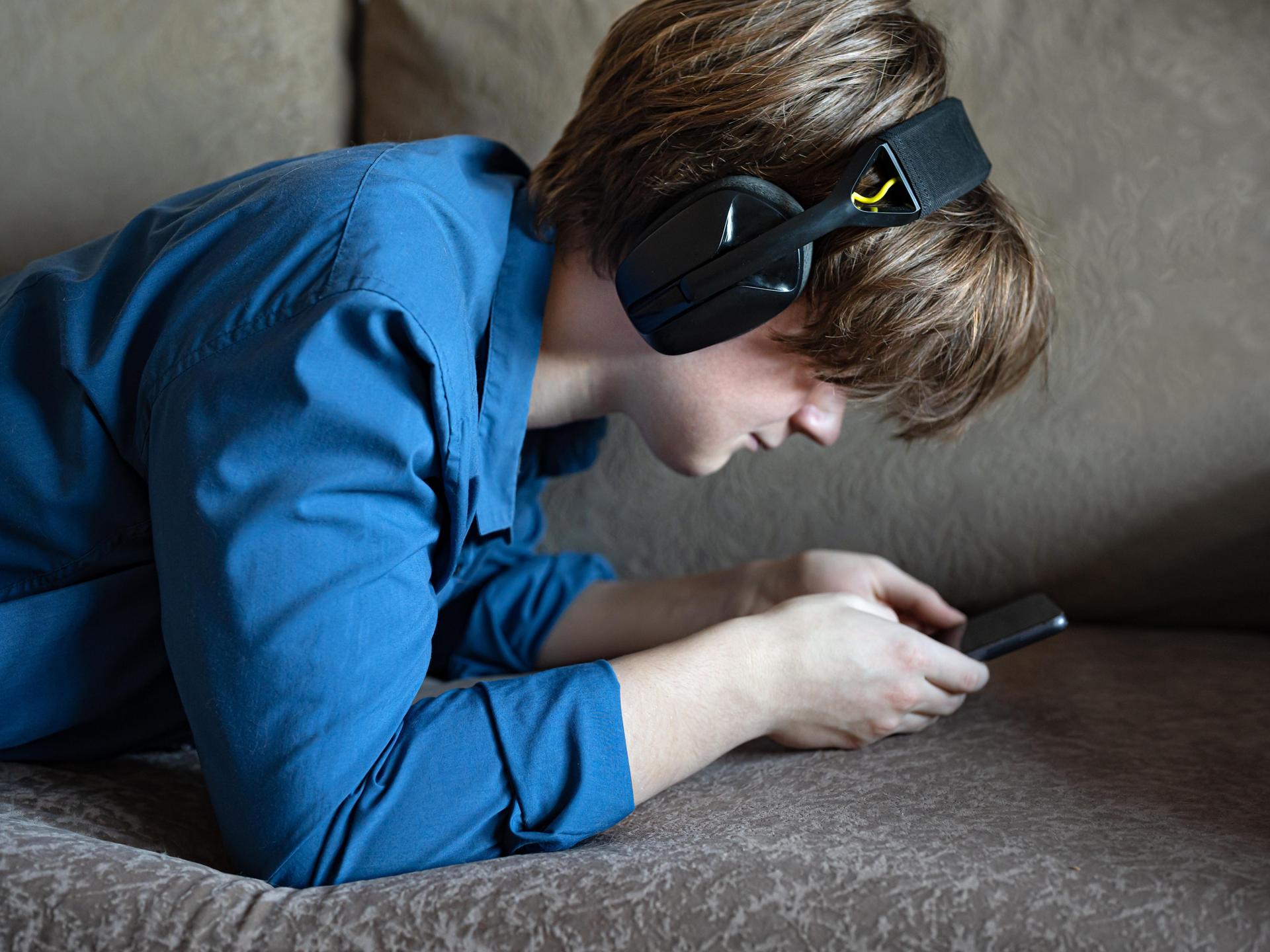 Ein Junge liegt mit seinem Smartphone auf einem Sofa. Er trägt Kopfhörer und starrt auf den Bildschirm seines Handys.