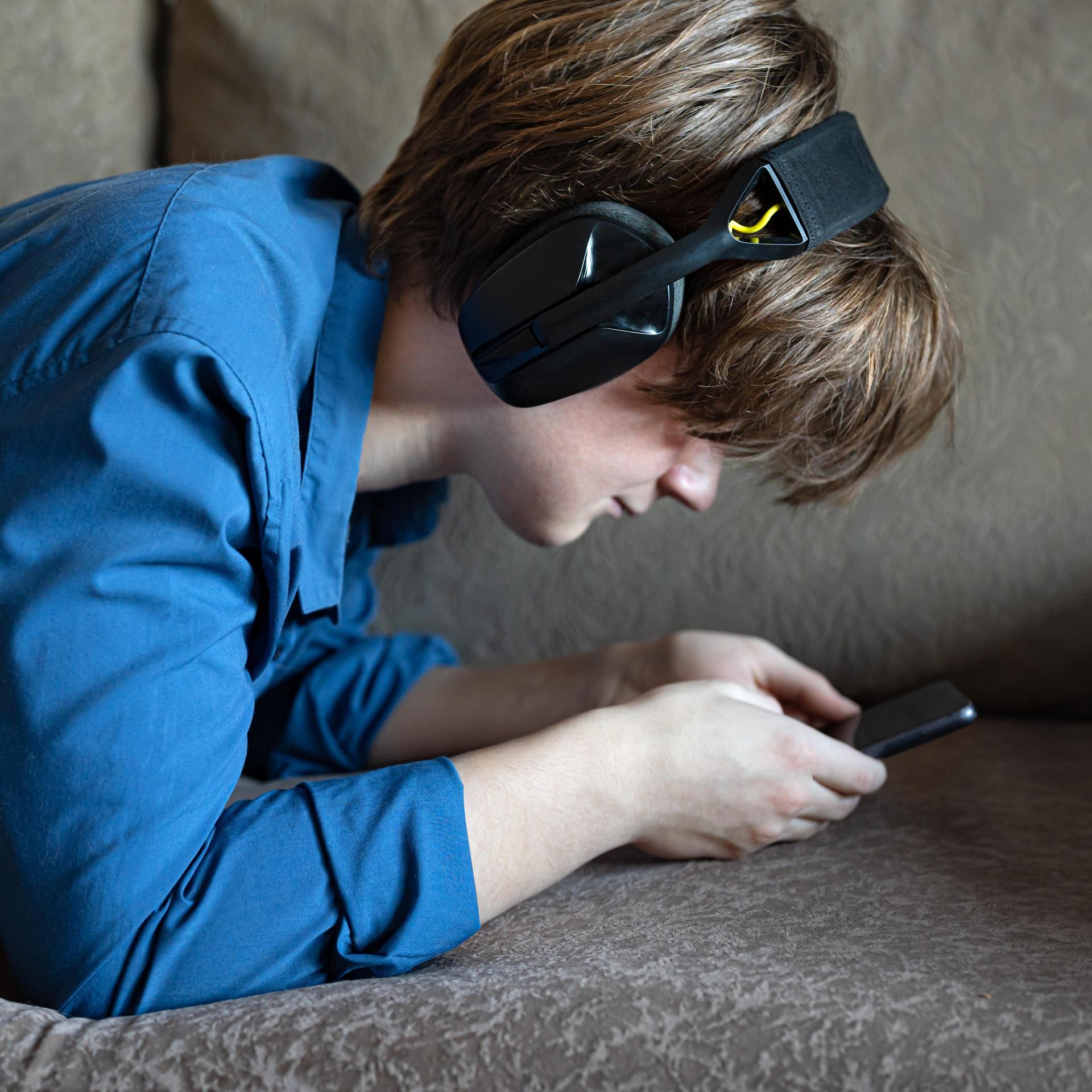 Ein Junge liegt mit seinem Smartphone auf einem Sofa. Er trägt Kopfhörer und starrt auf den Bildschirm seines Handys.