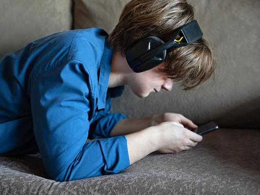 Ein Junge liegt mit seinem Smartphone auf einem Sofa. Er trägt Kopfhörer und starrt auf den Bildschirm seines Handys.