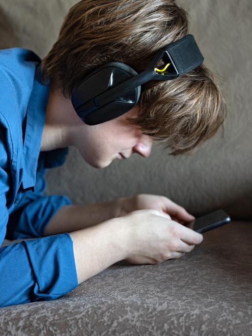 Ein Junge liegt mit seinem Smartphone auf einem Sofa. Er trägt Kopfhörer und starrt auf den Bildschirm seines Handys.