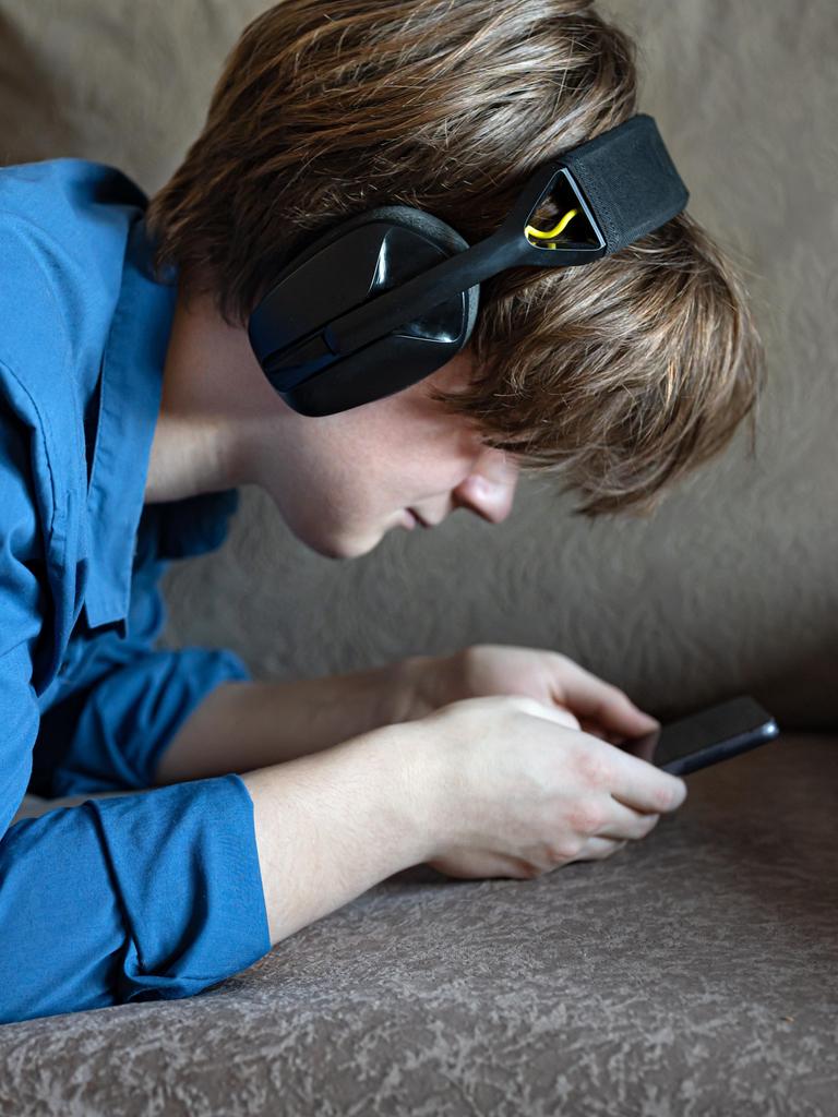 Ein Junge liegt mit seinem Smartphone auf einem Sofa. Er trägt Kopfhörer und starrt auf den Bildschirm seines Handys.