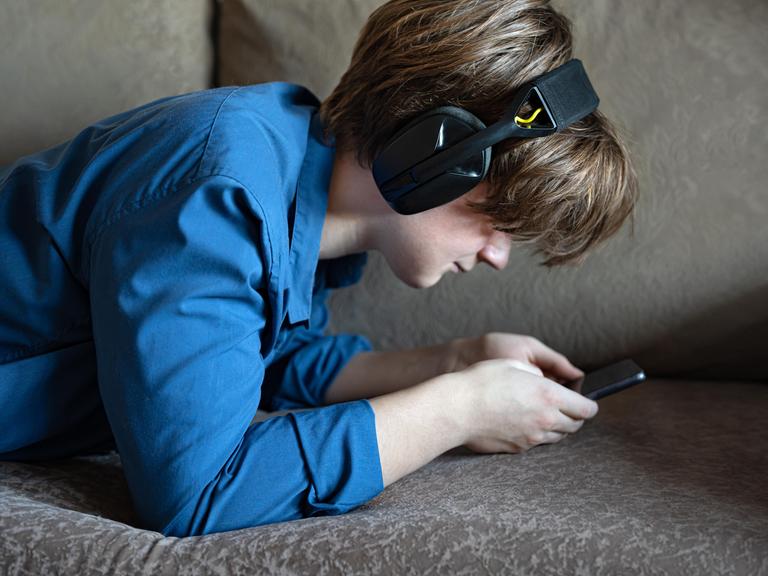 Ein Junge liegt mit seinem Smartphone auf einem Sofa. Er trägt Kopfhörer und starrt auf den Bildschirm seines Handys.